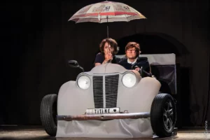 un couple dans une deux chevaux décapotée, l'une tient un parapluie, l'autre conduit