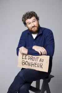 Olivier de Benoist, assis le bras croisé au dessus d'un carton où est écrit "Le droit au bonheur"