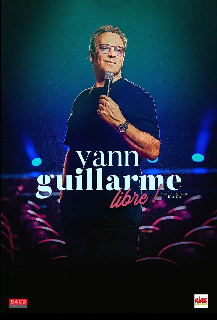Yann Guillarme debout, un micro à la main, au milieu d'une salle de spectacle, devant une scène en lumière.
