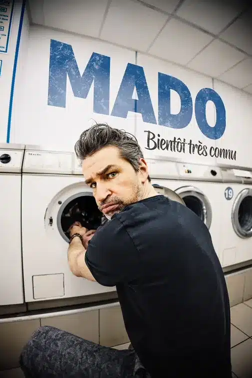 Affiche de MAdo dans Bientôt très connu Il est accroupi, en train de remplir une machine à laver dans un lavomatic, l'ai bougon