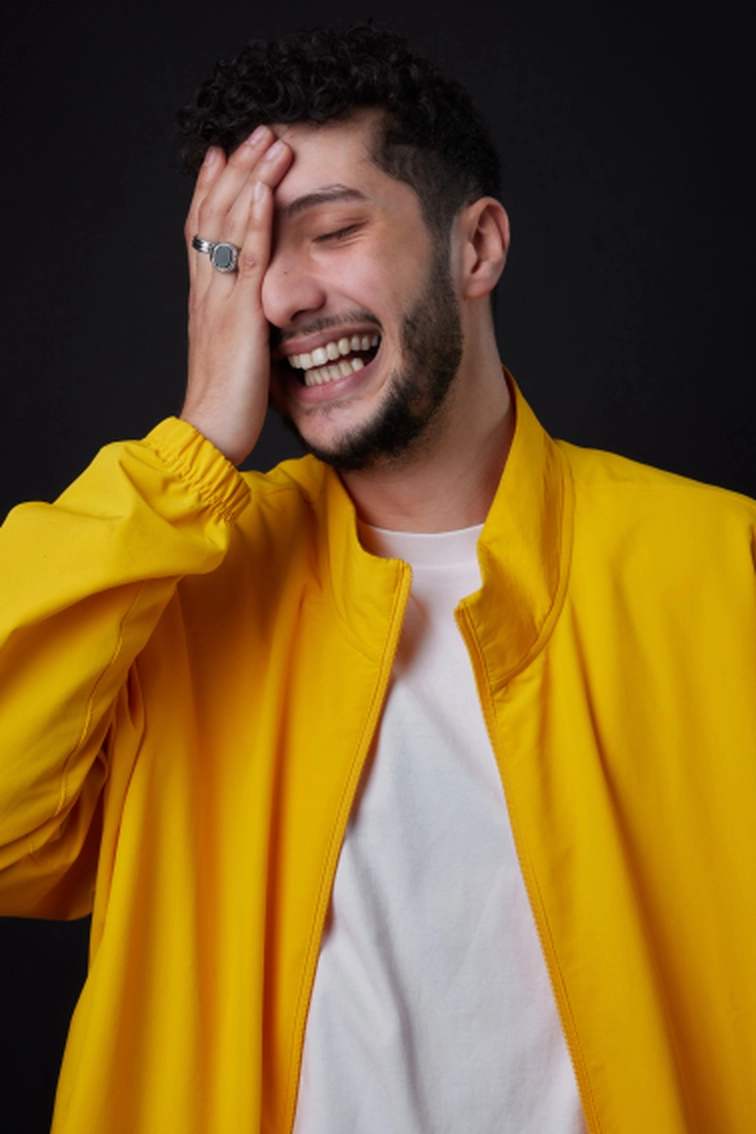 Portrait d'Elies Zoghlami souriant, chemise blanche blazer jaune, main devant un eil il rit