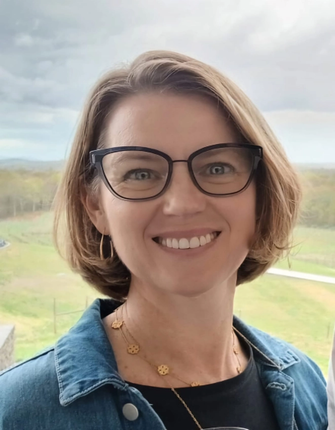 Kelly SMITH-cours atelier feldenkrais-web Portrait de Kelly Smith avec un grand sourire et des lunettes sur un fond bucolique