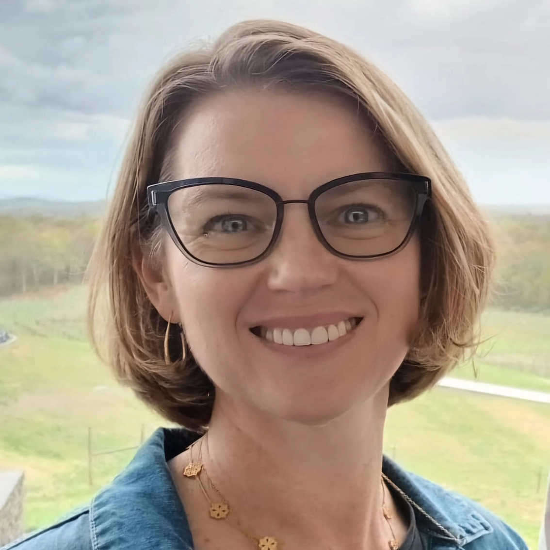 Portrait de Kelly Smith avec un grand sourire et des lunettes sur un fond bucolique