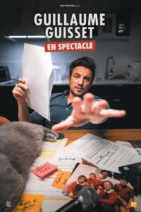 Photo de Guillaume Guisset assis à un bureau, un texte dans la main gauche, l'autre dirigée vers la caméra