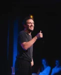 Photo de Guillaume Guisset dans son seul en scène, positif, un pouce en l'air, tee-shirt noir